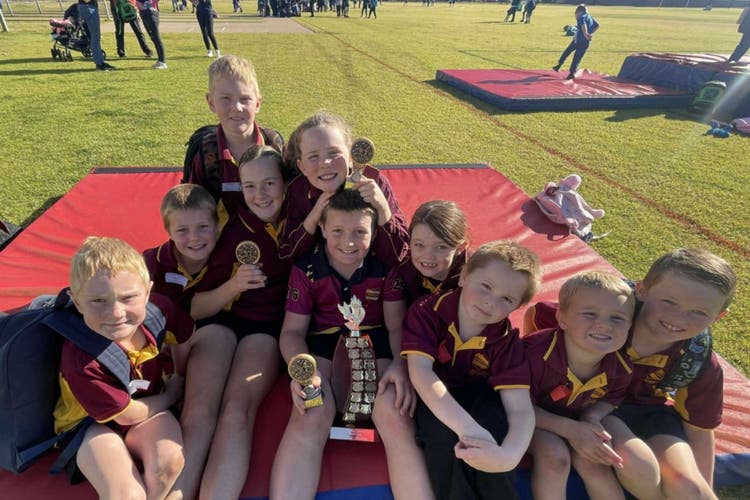 Students sitting on high jump mat holding trophies, smiling being happy